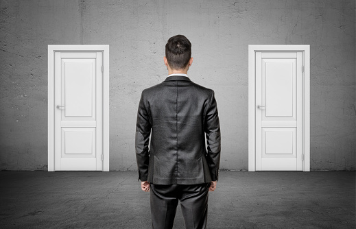 A businessman with his back turned stands between two identical closed white doors. Make right choice. Important decision. Never fail.