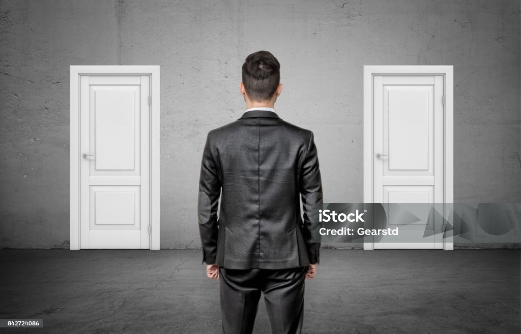 A businessman with his back turned stands between two identical closed white doors A businessman with his back turned stands between two identical closed white doors. Make right choice. Important decision. Never fail. Choice Stock Photo