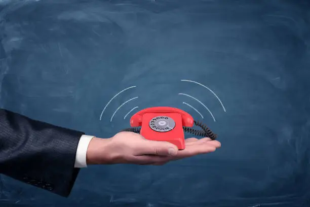 A businessman's palm hold a small red retro dial phone with lines showing soundwave emissions. Important call. Free number. Emergency connection.