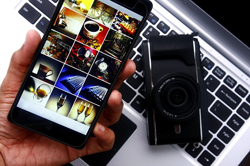 Photo of a hand holding a smartphone over a digital mirrorless camera and laptop computer