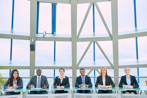 Group of smiling confident business specialists sitting in a row at one table and looking at camera in modern conference room with glassy roof