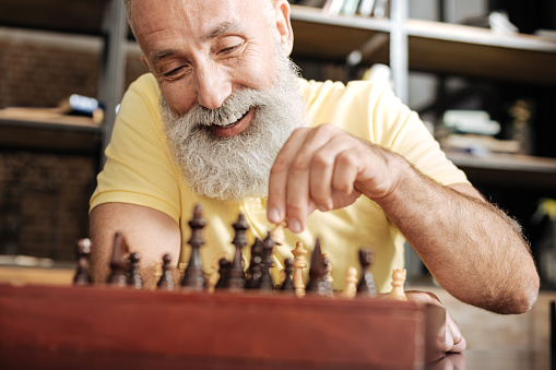 Brain game. Cheerful elderly man with a grey beard moving a pawn across a chessboard and smiling, obviously enjoying the game