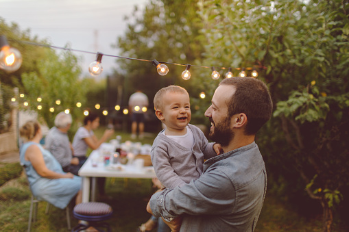 Photo of a multi-generation family having dinner outdoors in their back yard, while their little boy is celebrating his birthday