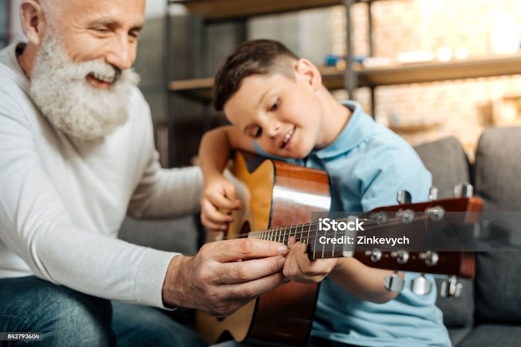 Smiling grandfather showing grandson how to play guitar Teaching with pleasure. Merry elderly man teaching his beloved grandson how to play guitar and strum chords while smiling happily Senior Adult Stock Photo