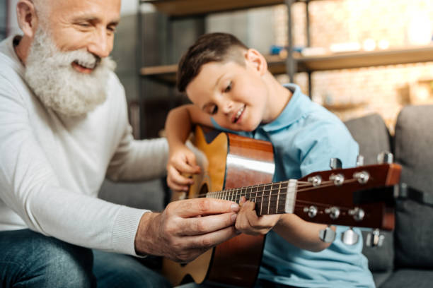 Smiling grandfather showing grandson how to play guitar Teaching with pleasure. Merry elderly man teaching his beloved grandson how to play guitar and strum chords while smiling happily chord stock pictures, royalty-free photos & images