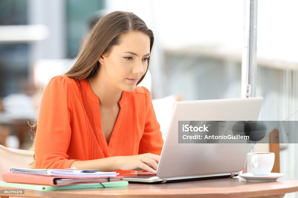 College student using a laptop in a coffee shop Single serious college student in orange using a laptop sitting in a coffee shop University Student Stock Photo