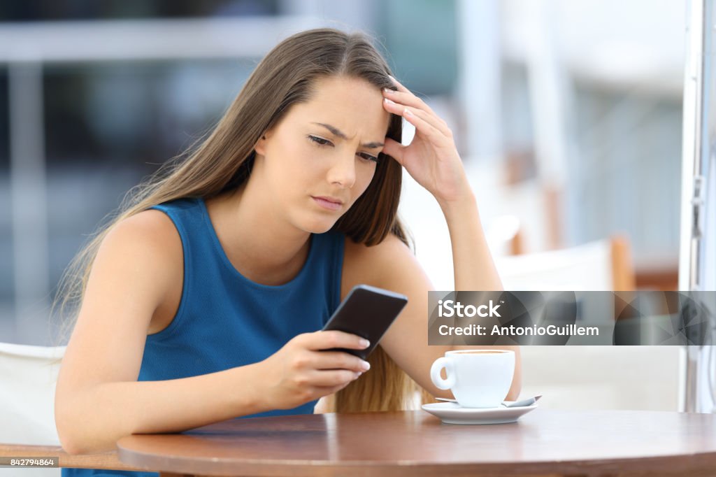 Worried woman holding phone and waiting for a call Single worried woman watching a mobile phone and waiting for a call or message sitting in a coffee shop Suspicion Stock Photo