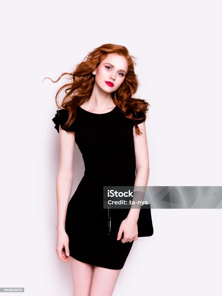 Beautiful young woman in luxury black cocktail dress holding clutch Portrait of young beautiful redhead woman with professional make-up looking gorgeous wearing luxury black dress and holding a clutch on white background Women Stock Photo