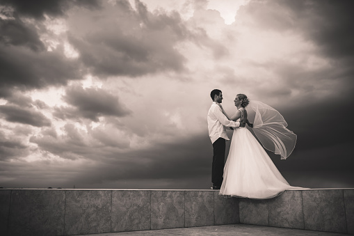 Loving bride and groom standing on the edge of a balcony and talking to each other against the sky. Copy space.