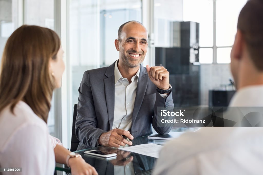 Happy financial agent smiling Mature financial agent in meeting sitting with young couple. Happy consultant talking with couple about their savings plan. Insurance advisor in a meeting with man and woman. Financial Advisor Stock Photo