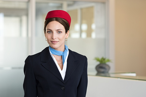 Portrait of young air hostess standing at airport and looking at camera. Portrait of flight assistant in uniform standing near check in counter. Happy agent wearing the hostess uniform at airport.