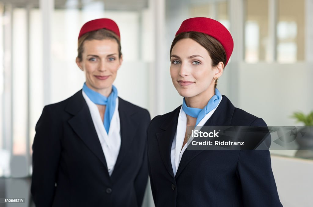 Smiling hostess at airport Two airhostess in uniform at airport looking at camera. Flight attendants standing together in blue and red uniform. Portrait of smiling young hostess ready for flight. Cabin Crew Stock Photo