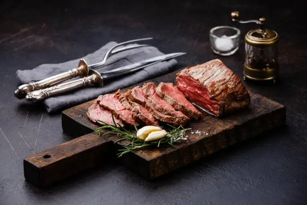 Sliced Tenderloin meat Roast beef on cutting board with knife and fork carving set, saltcellar and pepper mill