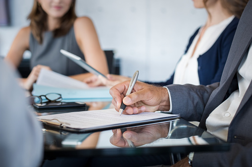 Closeup of hand of businessman holding pen and analyzing report during board meeting. Business people writing on document during annual meeting. Detail hand of business man fill the business contract.