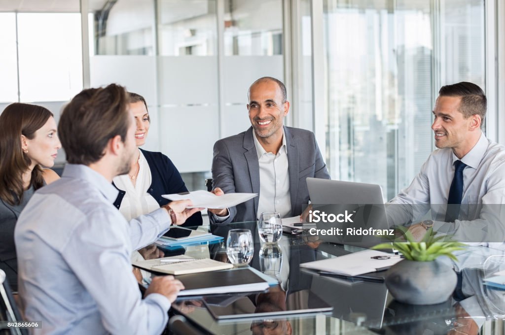 Businesspeople working in meeting Business man passing over documents to leader during meeting. Businessman passing necessary agreement to the business partner in conference room. Group of businessmen and businesswomen working together. Business Meeting Stock Photo