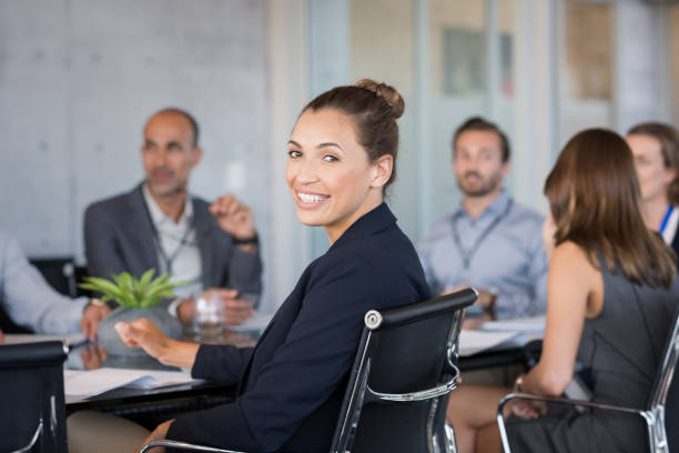 Young businesswoman attending meeting Young beautiful businesswoman with colleagues sitting in a modern board room. Proud smiling business woman sitting during a meeting and looking at camera. Portrait of a happy businesswoman with executives working. corporate boardroom stock pictures, royalty-free photos & images