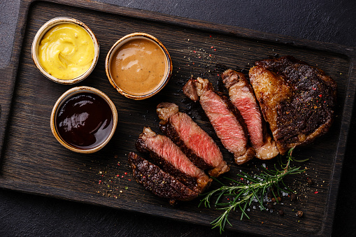 Sliced grilled Rib eye steak and three different sauces: Pepper sauce, Mustard and Barbecue on dark background close up