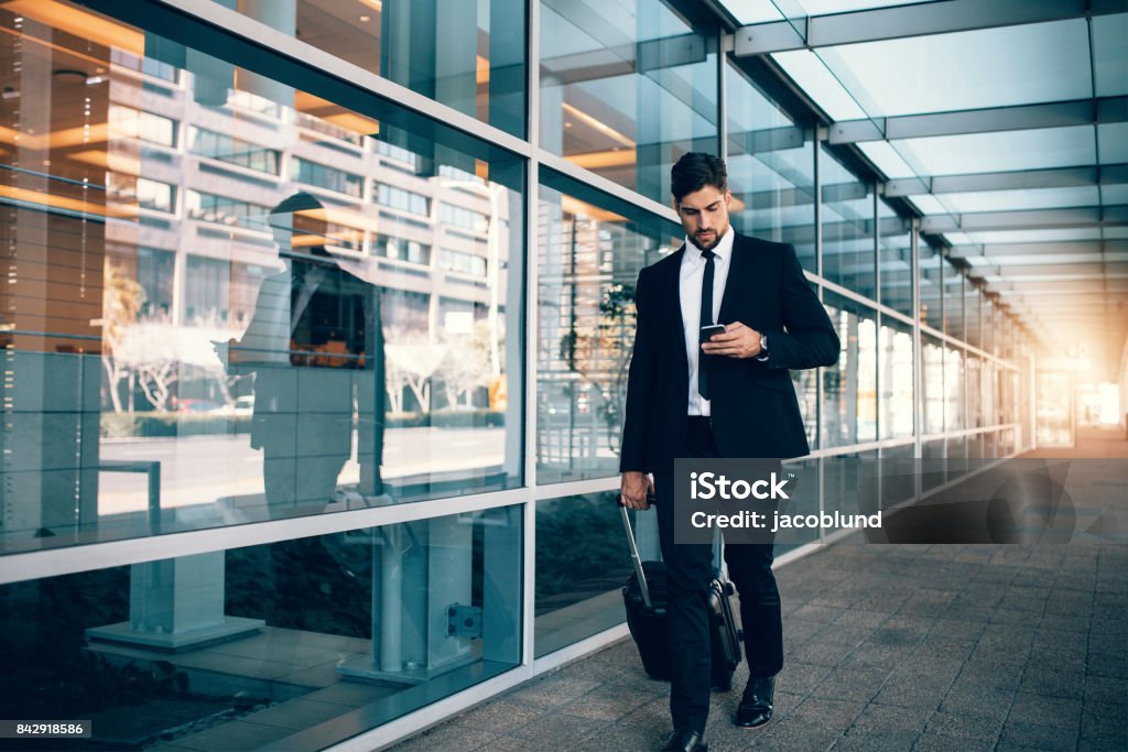 Businessman walking with luggage and using mobile phone at airport Businessman walking with luggage and using mobile phone at airport. Young man on business trip text messaging from his cell phone. Business Travel Stock Photo