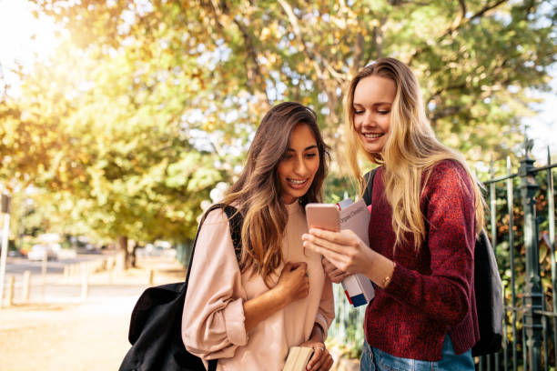 Female students using cellphone on road University female students reading message on smart phone outdoors. Beautiful students looking at mobile phone. education student mobile phone university stock pictures, royalty-free photos & images