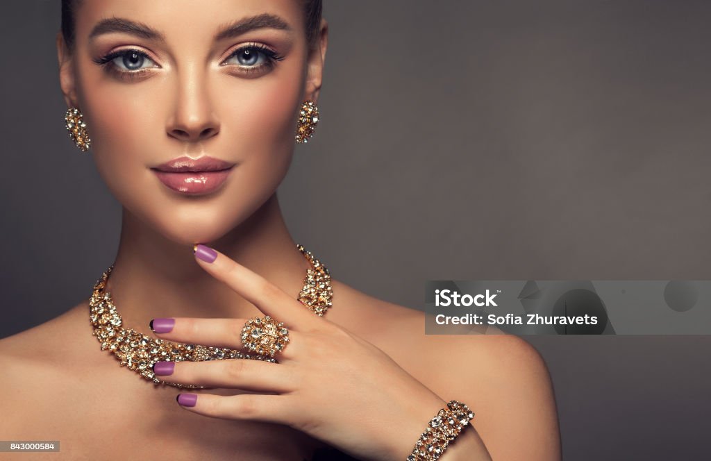 Magnificent lady in a perfect make up is shows jewelry set. Beauty portrait of young gorgeous woman is dressed in a jewelry set of necklace, ring and earings. Pretty  blue eyed model is demonstrating an attractive make up and manicure. Jewelry Stock Photo