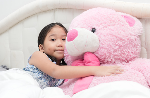 Portrait of expressive cute asian little girl hugging big pink bear, on bed in bedroom. Little girl playing with teddy bear