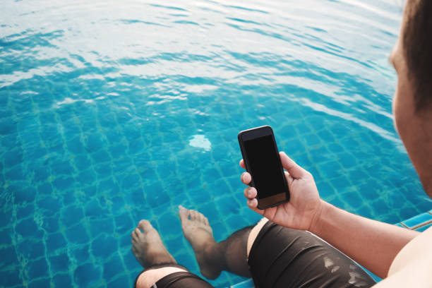 istock Close-up a man holding mobile smart phone with blank copy space on screen, with legs dipping in pool in summer for vacation. Clipping path on mobile phone screen 843131254