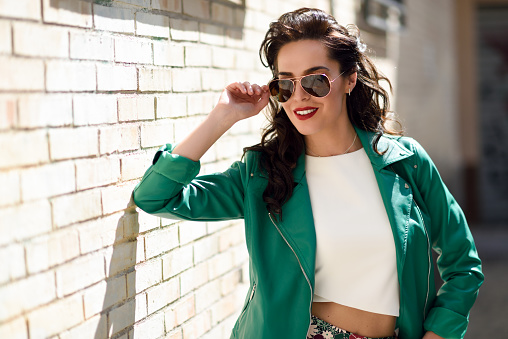 Young brunette woman with aviator sunglasses. Smiling girl, model of fashion, wearing green modern jacket, standing in urban background.
