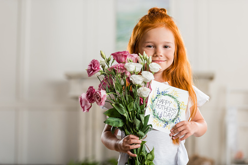 portrait of adorable smiling girl holding bouquet of flowers and happy mothers day greeting card