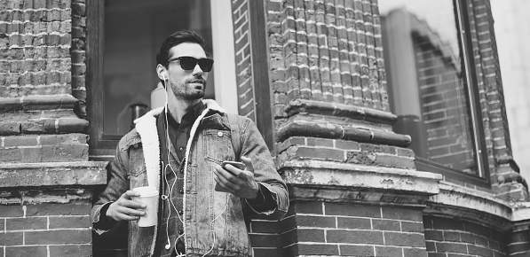 One man, standing on the city street, holding coffee cup, holding a mobile phone, listening music on headphones, black and white.