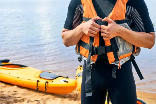 Young male hiker wearing wetsuit putting on a life vest with a yellow kayak on the blurred background