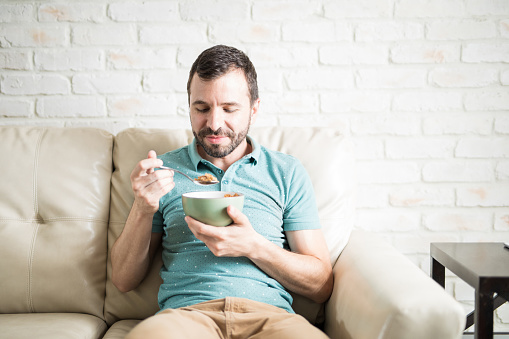Hispanic man having cereal for breakfast alone in his apartment sitting on the living room