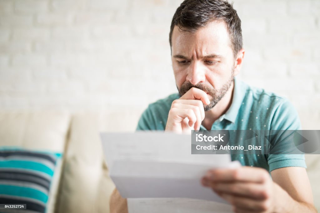 Man worried about his monthly payments Close up shot of a young adult man reading the mail in the living room and worried about his credit card payments Financial Bill Stock Photo