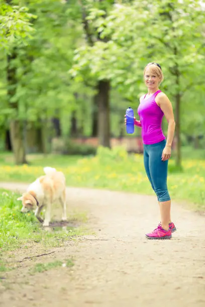 Woman running and walking in beautiful park with akita dog. Young girl exercising in bright forest outdoors. Power walking and drinking from water bottle in inspirational green woods landscape.