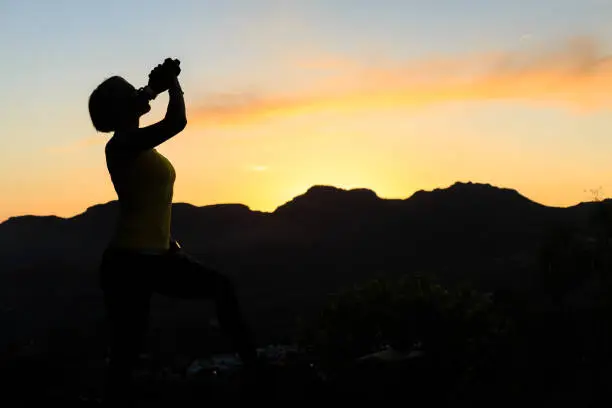 Woman hiking silhouette in mountains drinking water, sunset on mountain top. Female hiker, climber or trail runner with water bottle, looking at beautiful night sunset inspirational landscape.