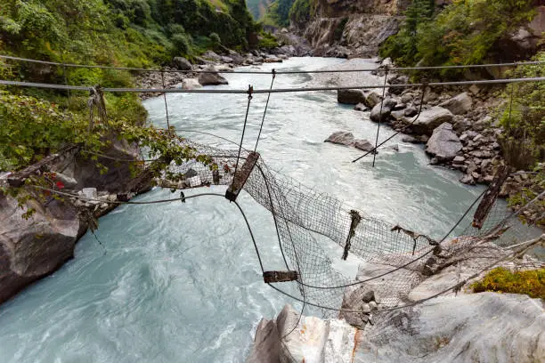Broken Suspension Bridge in Himalayas Nepal. Looking at river in canyon, forest and green trees. Annapurna Himal Range on Annapurna Circuit Trek. Autumn season in Nepal, Asia.