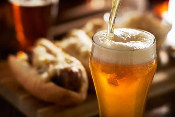 versando birra ambrata in tazza con bratwursts sullo sfondo - amber beer foto e immagini stock