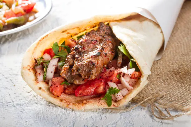 Photo of Turkish and Arabic Traditional Ramadan Adana Kebab Roll Wrap serving with yogurt, aubergine salad and hot pepper pickles on rustic wooden background.