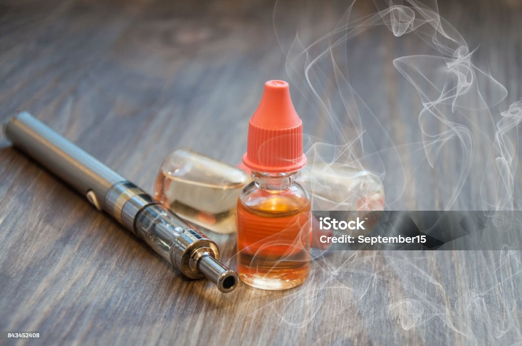 An electronic cigarette, a vape, and several bottles of liquid for refilling lie on a wooden background. with smoke around Arts Culture and Entertainment Stock Photo