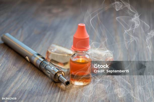 An Electronic Cigarette A Vape And Several Bottles Of Liquid For Refilling Lie On A Wooden Background With Smoke Around Stock Photo - Download Image Now