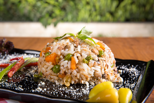 Brown Rice Risotto with baked pumpkin, boiled green pea, sliced tomato, green pickles, raw pepper and dried thyme for decoration black serving plate on brown burlap sack table cloth.