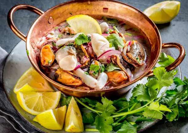 Peruvian Latin American seafood shellfish ceviche cebiche. Raw seafoods - mussels, shrimps, clams, squides marinated in lemon juice with red onion and coriander in cooper bowl, gray slate background.