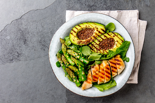 Healthy grilled chicken, grilled avocado and asparagus salad with linen seeds. Balanced lunch in bowl. Gray slate background. Top view.