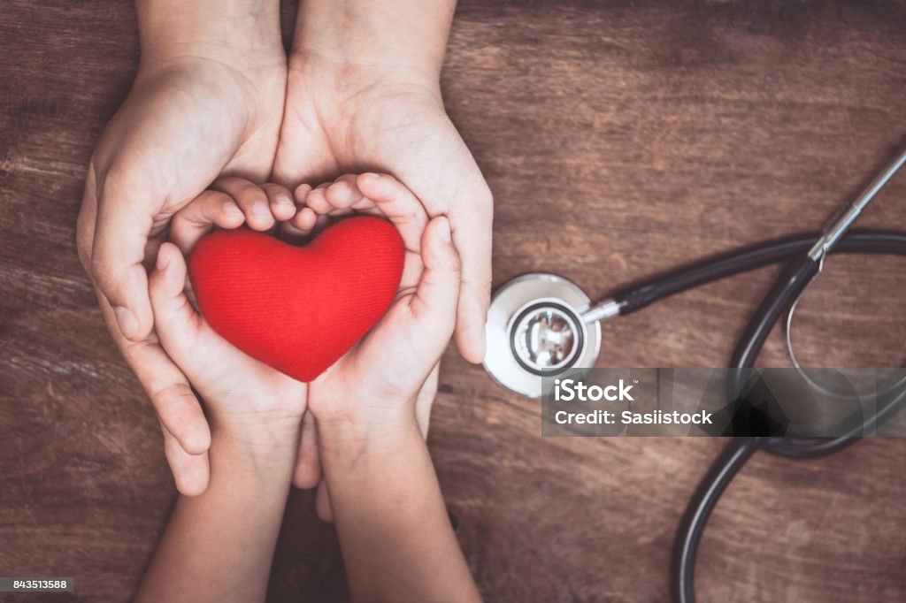 Corazón rojo en manos de la mujer y el niño y con el estetoscopio del médico sobre fondo de madera - Foto de stock de Niño libre de derechos