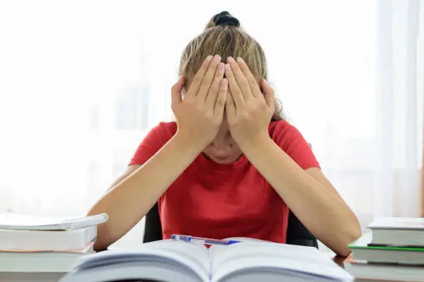 Schoolgirl holds her head in her hands over a pile of books on the desk and is tired and frustrated with the homework problem at school.