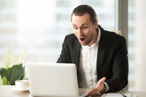 Frustrated young businessman looks at laptop screen screaming in anger at workplace in office. Unexpected high bill, unpaid debt, failing financial report, tax delinquency, breach of contract concept.