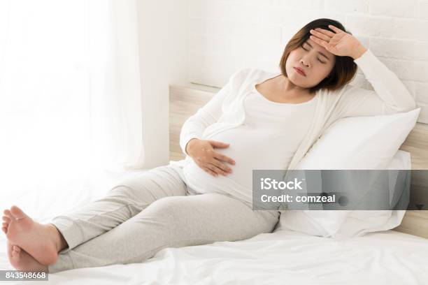 Asian Pregnant Woman With Hand On Forehead Suffering Headache Sitting On Bed At Home Stock Photo - Download Image Now
