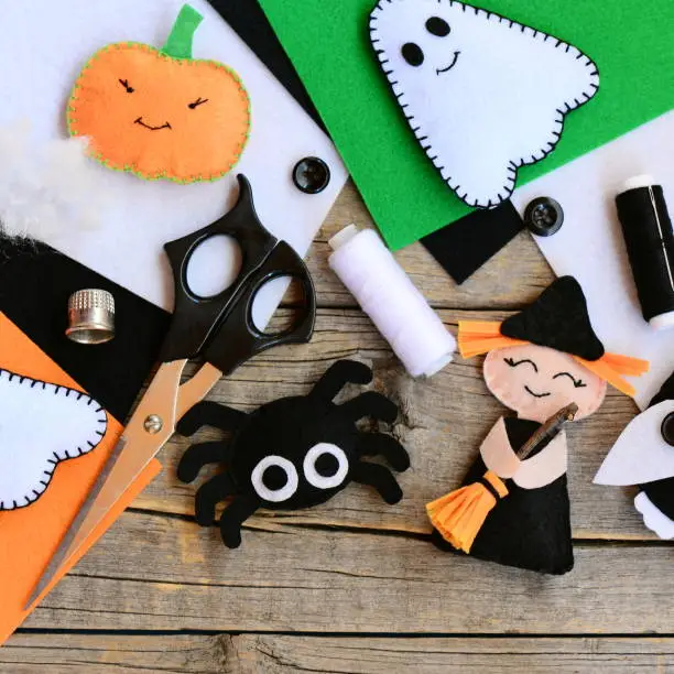 Photo of Felt toys for Halloween home decor clearance. Felt witch with broom, pumpkin head, ghost, spider. Handmade Halloween decorations. Felt sheets, scissors, threads on a wooden table. Top view. Closeup