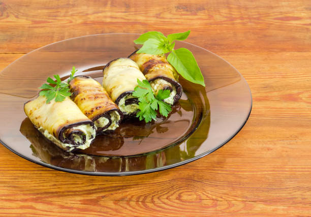 Fragment of a dark glass dish with several eggplant rolls stuffed with tuna and processed cheese and decorated with parsley and basil twigs on a surface of an old wooden planks
