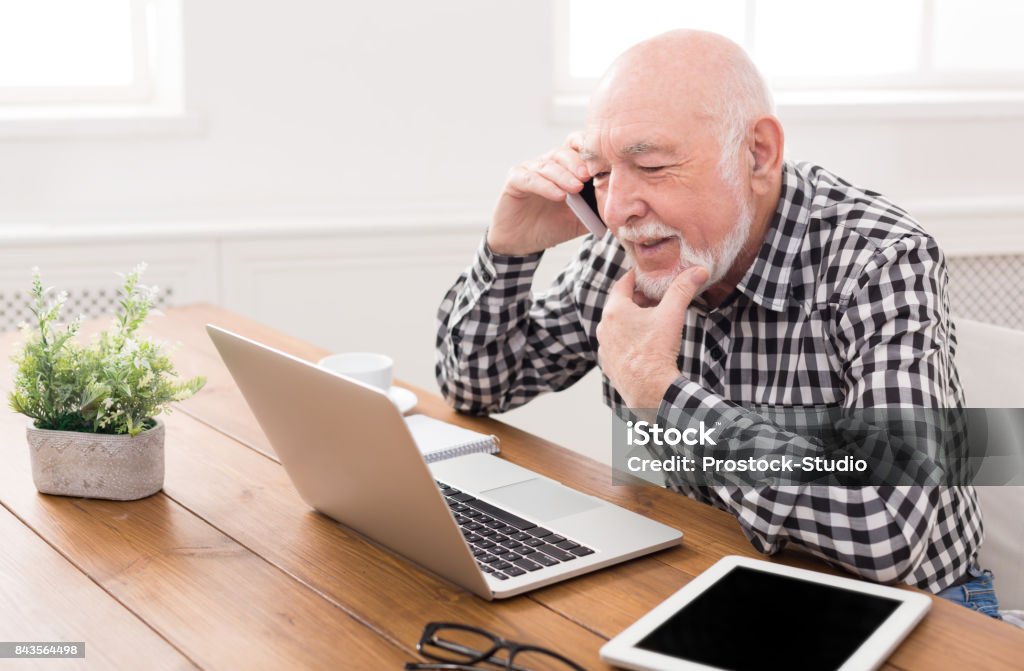 Senior man talking on phone with laptop Senior man talking on phone and using laptop, sitting at table at home. Modern technology, communication concept Senior Men Stock Photo