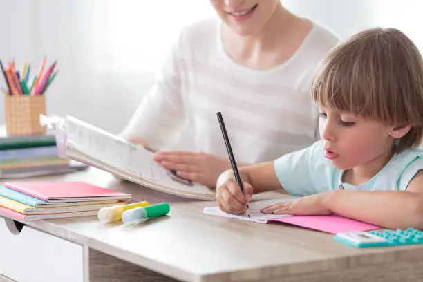 Boy focusing on homework while sitting with mother at desk with notebook and colored pens